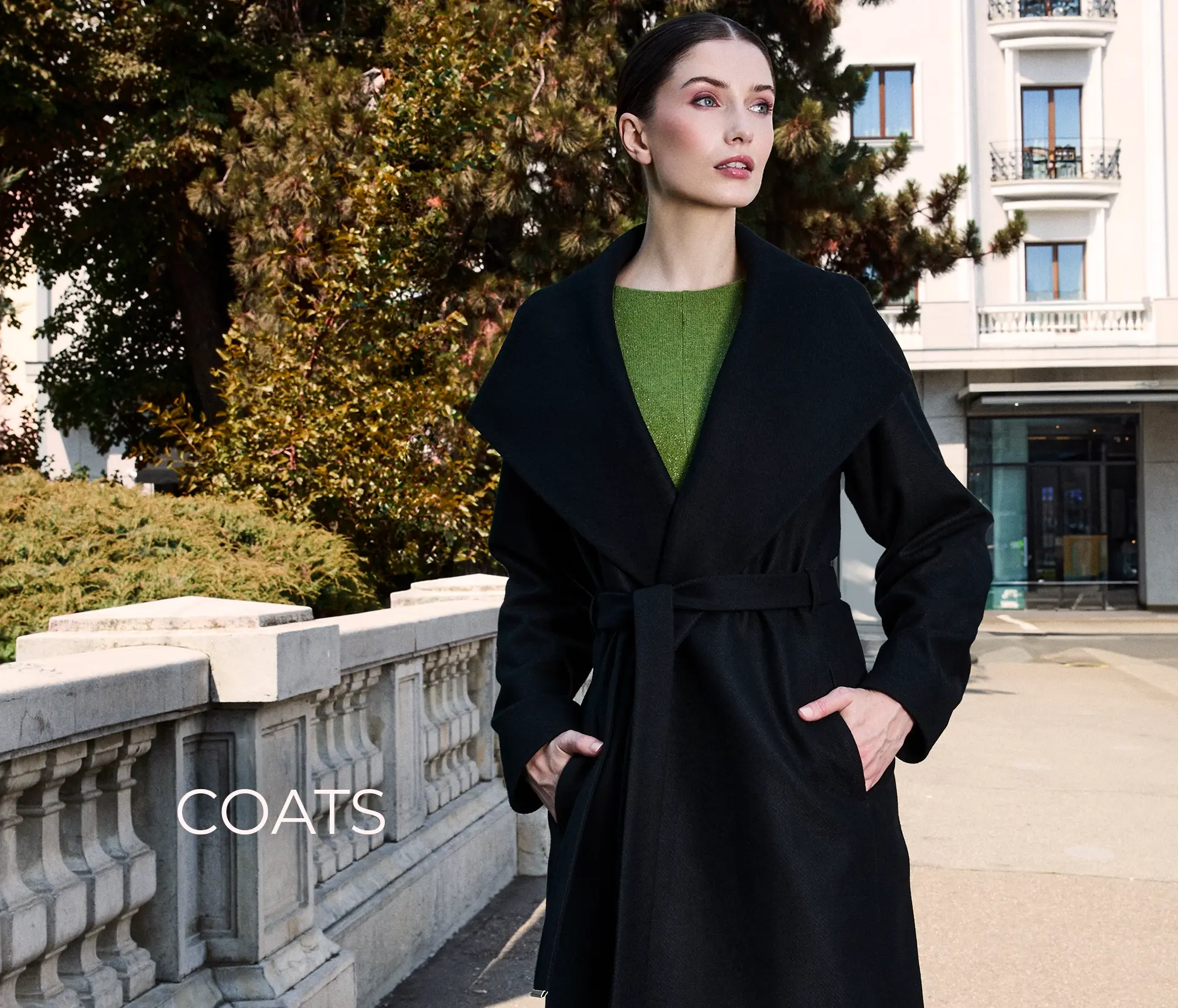 Woman in a black coat with a green sweater, standing outdoors, looking elegant. Dika women's fashion.